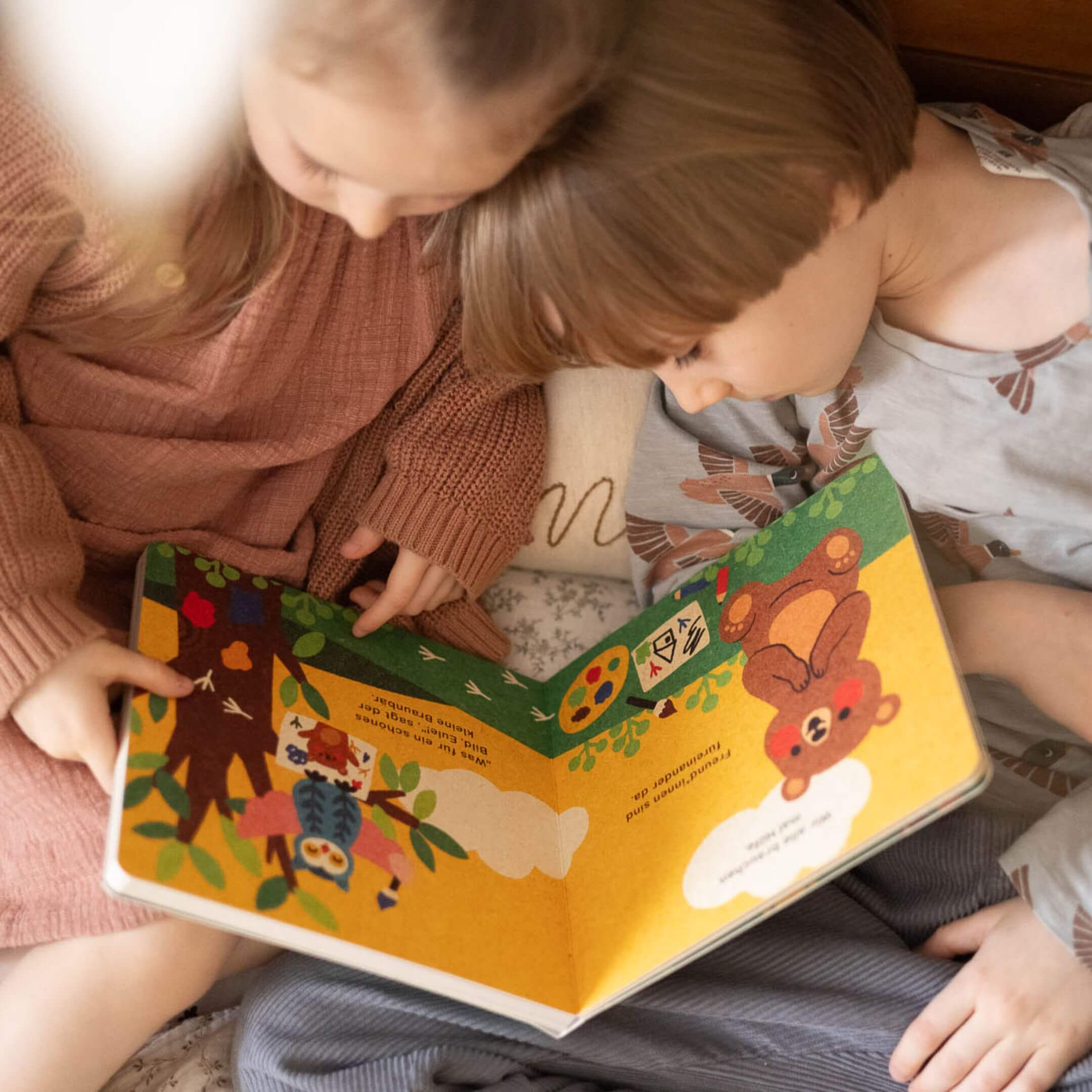 Zwei kleine Kinder sitzen zusammen und lesen "Hilfe brauchen ist ok" von Annelies Draws. Während sie sich auf die farbenfrohe Geschichte konzentrieren, spiegelt ihr gemeinsamer Moment auf wunderbare Weise Freundschaft und Empathie wider.