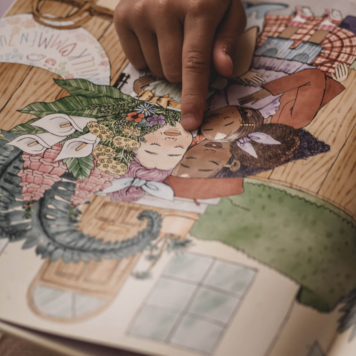 Die Hand eines Kindes zeigt auf eine Illustration in "Mama und Mami und ich: Die große Vermissung" von Nina LaCour, die zwei Menschen mit geschlossenen Augen zwischen Blumen in einem gemütlichen Zimmer zeigt - eine sanfte Szene über das Fehlen eines Elternteils während der Trennung.