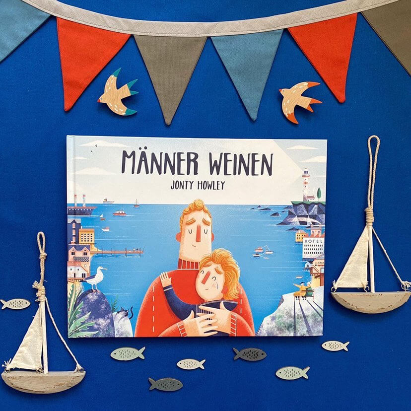Das Kinderbuch "Männer weinen" von Jonty Howley liegt auf blauem Hintergrund, umgeben von bunter Wimpelkette sowie maritimen Dekorationen - ein Buch über Emotionen bei Männern.