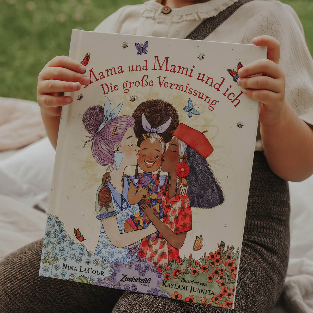 Ein Kind hält Mama und Mami und ich: Die große Vermissung von Nina LaCour, in der drei Frauen ein Kind inmitten von Blumen und Schmetterlingen umarmen. Die Geschichte thematisiert behutsam Trennungsphasen und das Vermissen eines Elternteils.