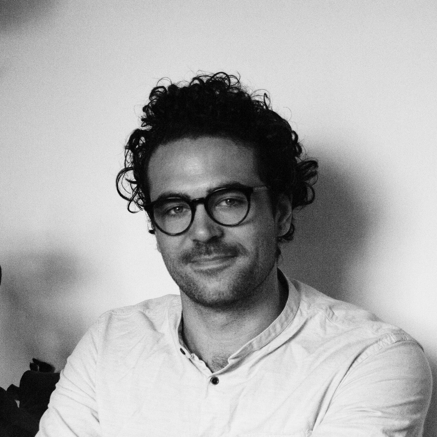 Das Schwarz-Weiß-Foto eines Mannes mit lockigem Haar und Brille in einem hellen Button-up, der sitzend und sanft in die Kamera lächelt, erinnert an die stille Emotionalität in Jonty Howleys "Männer weinen".